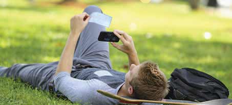 man laying in the grass with a skateboard as he takes a photo of a check with his mobile device